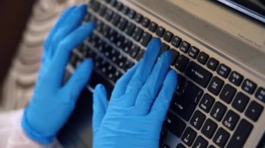 Yakından tanınmayan mavi koruyucu eldivenli sağlıkçı bilgisayarda uzaktan çalışıyor. Doktor elleri klavyeye yazı yazarken üst görüntüyü kapatın. Bilgisayar kullanan bir adam. Pandemik dönemde koruma kavramı.