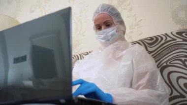 Genç hemşire beyaz koruyucu elbise, tıbbi maske, mavi eldiven giyer, dizüstü bilgisayar kullanır. Kadın hekim oturma odasında uzaktan kumandayla çalışıyor. COVID-19 'dan Sosyal Mesafe Kapatma Salgını