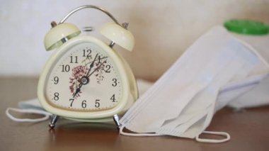 Beyaz koruyucu tıbbi maskelerin yakınındaki ahşap masada çalan bej retro alarm saati. Sabah 7 'de. Koronavirüs, nam-ı diğer kovid-19 virüsü tehdidi nedeniyle karantinaya alınmıştır. Kilitleyin. Kapalı