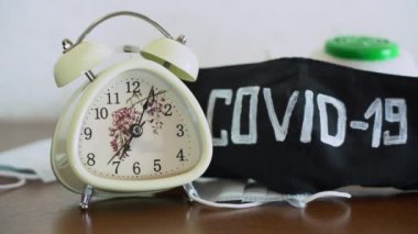Siyah koruyucu yüz maskelerinin yanında kahverengi ahşap masada, üzerinde COVID-19 yazan beyaz çalar saat çalıyor. Saat 7 'de Coronavirus covid-19 virüsü tehdidiyle karantinaya alınmış. Kilitle.