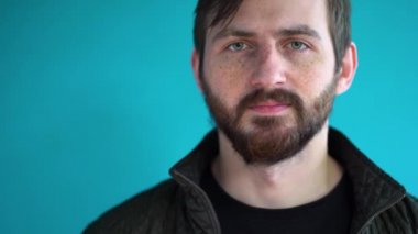 Sakallı, beyaz tenli, çilli bir adamın portresi. Siyah ceketli, kısa kahverengi saçlı çekici bir erkek mavi duvar arka planına karşı koruyucu bir yüz maskesi takıyor ve çıkarıyor..