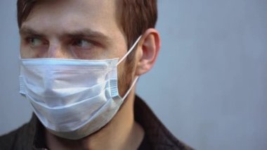 Hasta, sakallı, yeşil gözlü, koruyucu, tek kullanımlık tıbbi yüz maskesi takan ve kameraya uzun uzun bakan hasta bir adam. Gri arka planda hasta ve güzel bir adamın portresi. İzolasyon kavramı.