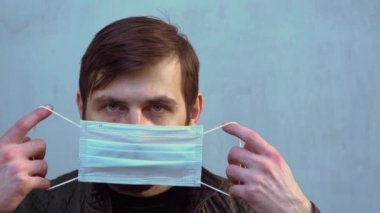 Koyu renk ceketli kısa kahverengi saçlı sakallı adam koruyucu bir tek kullanımlık turkuaz maske takıyor. Dışarıdaki gri duvarlı arka planda ağır çekimde kameraya bakıyor.