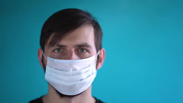 青い壁の背景に隔離されたカメラをまっすぐに見て 白い保護医療面マスクを身に着けている緑の目を持つ美しい自信の若い男 コロナウイルスの脅威の概念を停止します 自己分離 — ストック動画