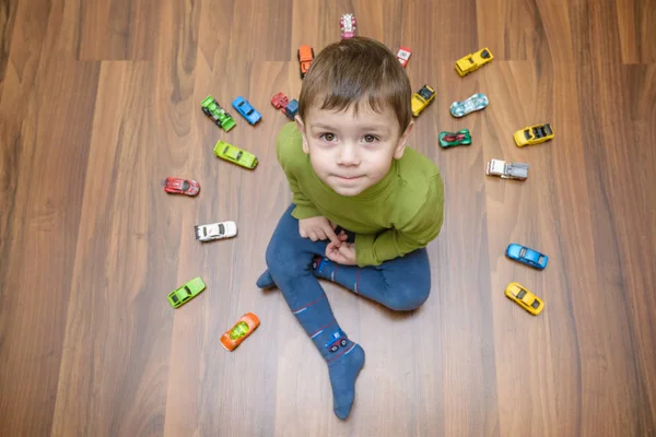 Pequeño niño caucásico jugando con un montón de coches de juguete en interiores. Niño vistiendo camisa verde. Feliz preescolar divirtiéndose en casa o guardería — Foto de Stock