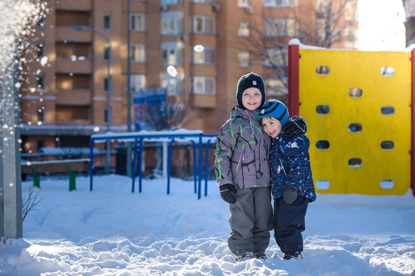 Два маленьких мальчика в разноцветной одежде играют на улице во время снегопада. Активный отдых с детьми зимой в холодные дни. Счастливые братья и сестры и близняшки веселятся на снегу — стоковое фото