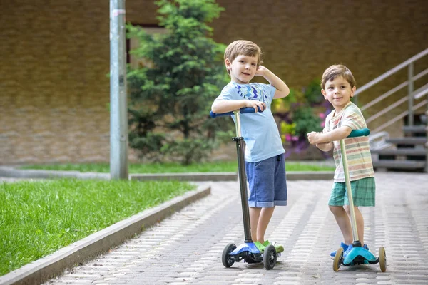 Dois meninos bonitos, competir em scooters equitação, ao ar livre no parque, verão . — Fotografia de Stock