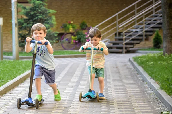 Dois meninos bonitos, competir em scooters equitação, ao ar livre no parque, verão . — Fotografia de Stock