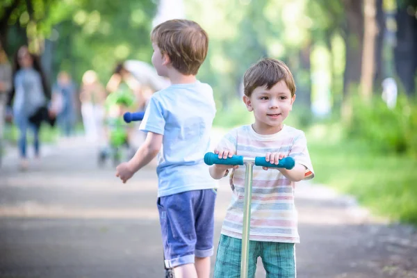 Two cute boys, compete in riding scooters, outdoor in the park, summertime. — Stock Photo, Image