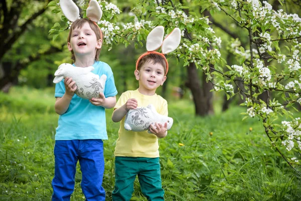 Two little kids boys and friends in Easter bunny ears during traditional egg hunt in spring garden, outdoors. Siblings having fun with finding colorful eggs. Old christian catholoc tradition — Stock Photo, Image