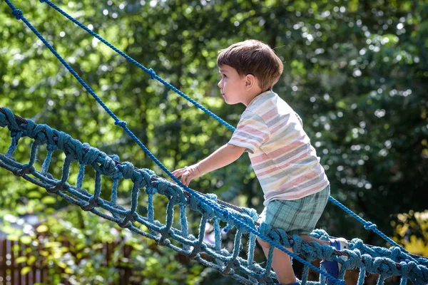 Little climber takes the rope bridge. Boy has fun time, kid climbing on sunny warm summer day — Stock Photo, Image