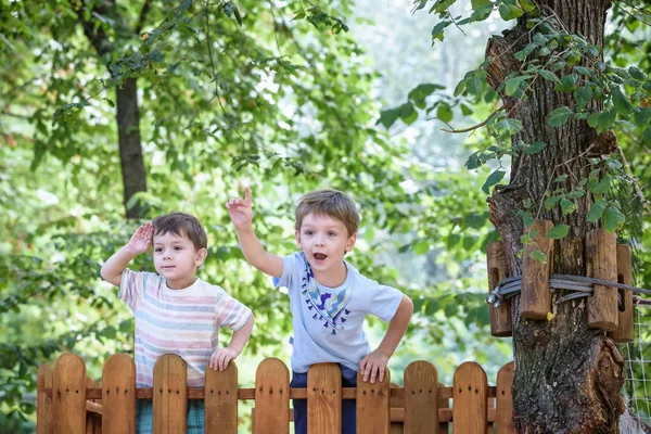 Dos hermanitos jugando juntos de pie en un parque de madera en el parque de verano. El chico le muestra algo interesante a su hermano. Niño mirando a través de un tubo imaginario — Foto de Stock