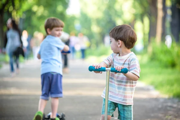 Two cute boys, compete in riding scooters, outdoor in the park, summertime. — Stock Photo, Image