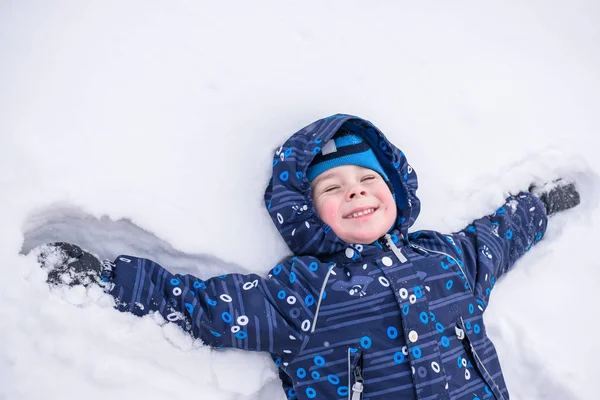 Ragazzino carino in abiti invernali colorati facendo angelo della neve, sdraiato su. Tempo libero attivo all'aperto con i bambini in. Bambino felice. . — Foto Stock