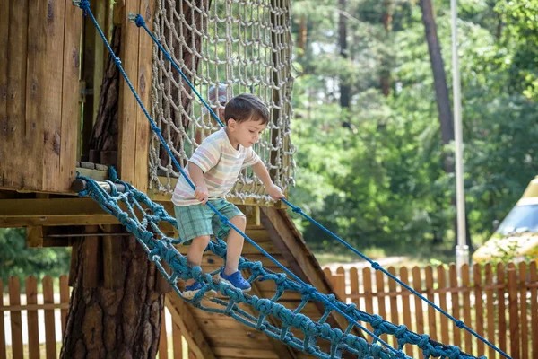 O pequeno alpinista fica com a ponte da corda. Menino se diverte, criança escalando no dia quente ensolarado do verão — Fotografia de Stock