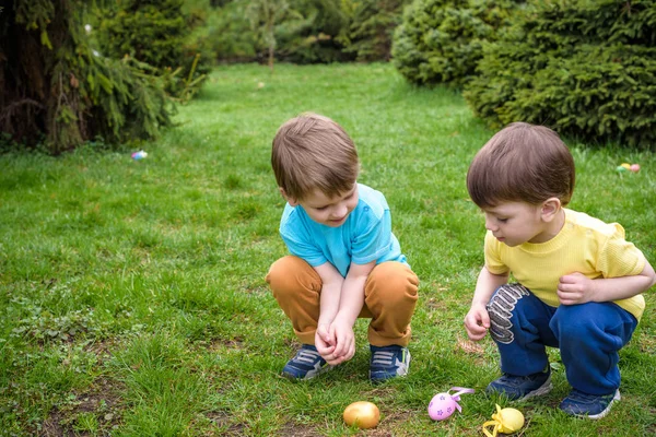 Los niños en la búsqueda de huevos de Pascua en el floreciente jardín de primavera. Niños en busca de huevos de colores en el prado de flores. niño pequeño y su hermano amigo niño jugar al aire libre — Foto de Stock