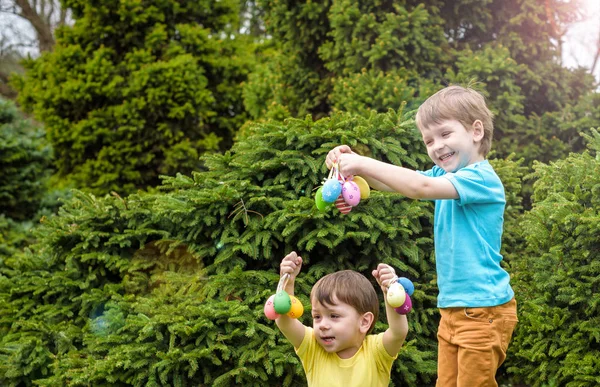 Los niños en la búsqueda de huevos de Pascua en el floreciente jardín de primavera. Niños en busca de huevos de colores en el prado de flores. niño pequeño y su hermano amigo niño jugar al aire libre —  Fotos de Stock