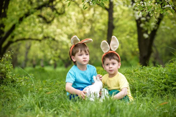 Two little kids boys and friends in Easter bunny ears during traditional egg hunt in spring garden, outdoors. Siblings having fun with finding colorful eggs. Old christian catholoc tradition — Stock Photo, Image