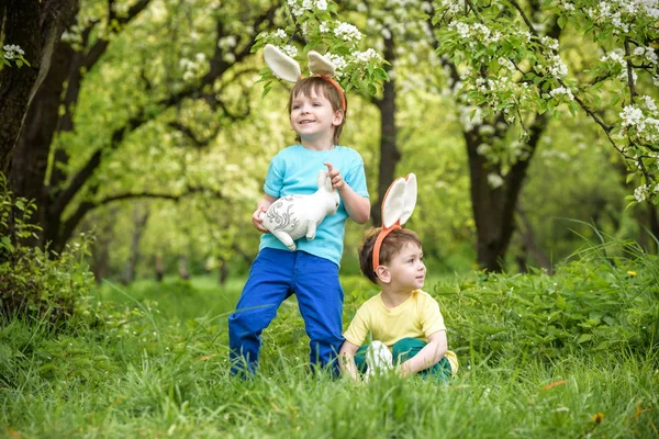 Two little kids boys and friends in Easter bunny ears during traditional egg hunt in spring garden, outdoors. Siblings having fun with finding colorful eggs. Old christian catholoc tradition — Stock Photo, Image