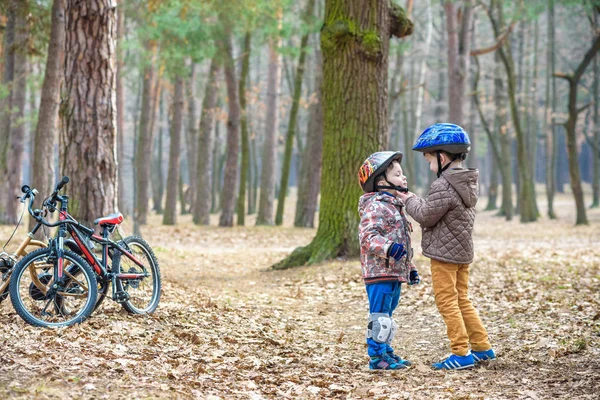 Dwa małe dzieci chłopców, najlepsi przyjaciele w lesie jesienią. Starszy brat pomaga młodsze dziecko umieścić jego kask rowerowy. Szczęśliwy rodzeństwo z rowerami. — Zdjęcie stockowe