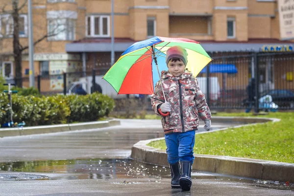 Mały chłopiec, grając w deszczowe lato park. Dziecko z rainbow kolorowy parasol, wodoszczelny płaszcz i buty skoki w kałuży i błoto w deszczu. Dziecko, chodzenie w jesień prysznic zabawy na zewnątrz przy każdej pogodzie — Zdjęcie stockowe