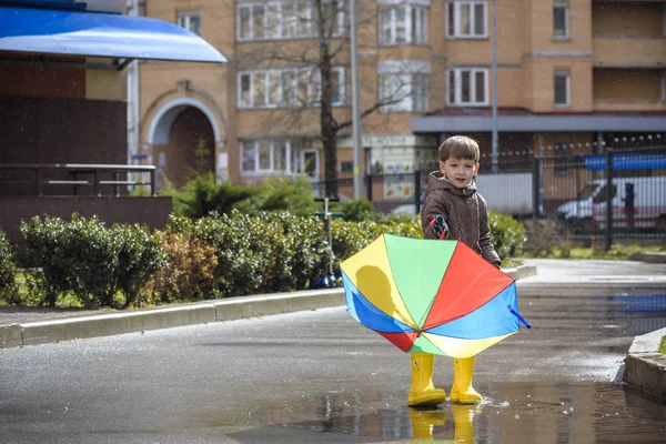 Mały chłopiec, grając w deszczowe lato park. Dziecko z rainbow kolorowy parasol, wodoszczelny płaszcz i buty skoki w kałuży i błoto w deszczu. Dziecko, chodzenie w jesień prysznic zabawy na zewnątrz przy każdej pogodzie — Zdjęcie stockowe