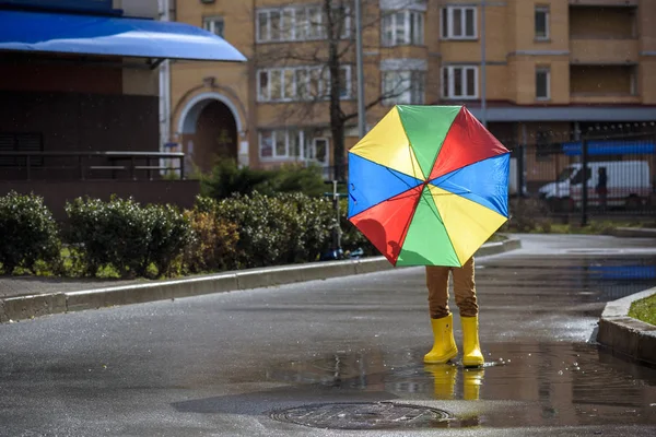 Мальчик играет в дождевом летнем парке. Ребенок с разноцветным радужным зонтиком, водонепроницаемым пальто и сапоги, прыгающие в луже и грязи под дождем. Ребенок ходит в осенний душ на открытом воздухе весело при любой погоде — стоковое фото