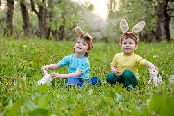 Two little kids boys and friends in Easter bunny ears during traditional egg hunt in spring garden, outdoors. Siblings having fun with finding colorful eggs. Old christian catholoc tradition — Stock Photo, Image