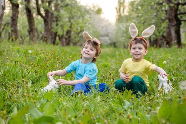 Dva malí kluci a přátelé na velikonočních králičích uších při tradičním lovu vajíček na jarní zahradě, venku. Sourozenci se baví hledáním barevných vajec. Stará křesťanská katolická tradice — Stock fotografie