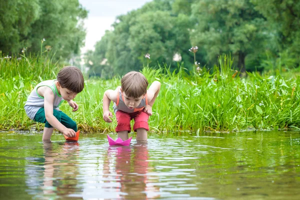 Dos hermanitos jugando con barcos de papel junto a un río en un cálido y soleado día de verano . — Foto de Stock