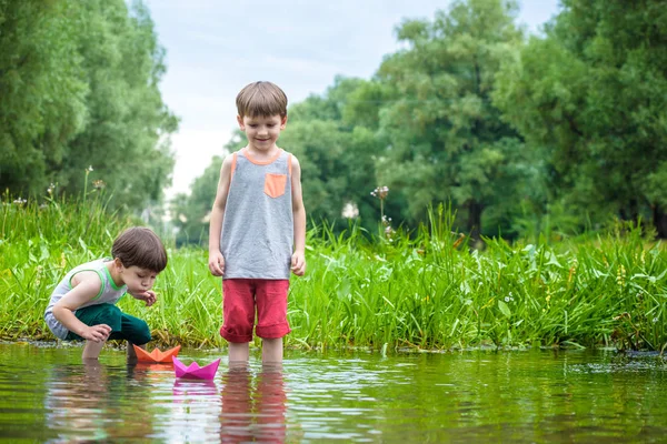 Dos hermanitos jugando con barcos de papel junto a un río en un cálido y soleado día de verano . — Foto de Stock