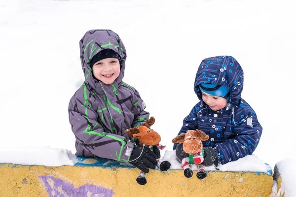 Two adorable preschool kids brother boys in winter wear sit amoung snow and play with toy reindeer — Stock Photo, Image