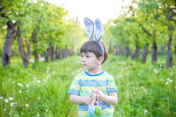 Killen på påskägget i blommande våren trädgård. pojke söker färgglada ägg i blomma äng — Stockfoto