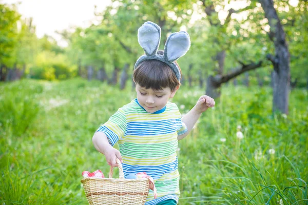 Веселый мальчик держит корзину с яркими пасхальными яйцами, стоящими на траве в парке после охоты за яйцами — стоковое фото
