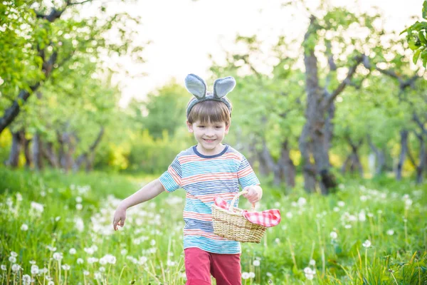 Веселый мальчик держит корзину с яркими пасхальными яйцами, стоящими на траве в парке после охоты за яйцами — стоковое фото