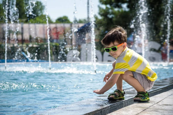 Niño juega en la plaza cerca de la piscina con chorros de agua en la fuente en el soleado día de verano. Ocio activo de verano para niños en la ciudad —  Fotos de Stock