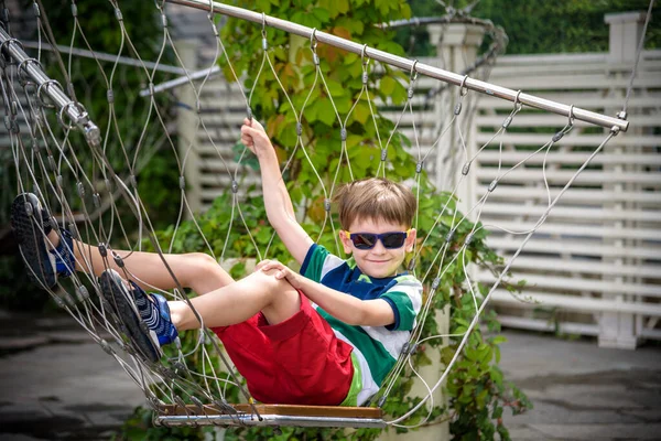 Little adorable kid sitting on chain swing during summer holidays on vacation. Boy relaxed and happy wearing casual clothes in sunglasses. Active summer leisure for kids in the city concept — Stock Photo, Image