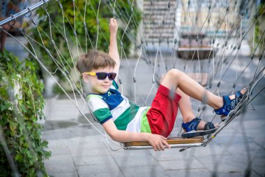 Yaz tatilinde zincirli salıncakta oturan sevimli çocuk. Çocuk rahat ve güneş gözlüğü takarak mutlu. Şehir konseptinde çocuklar için aktif yaz tatili.