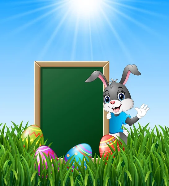 Conejito de dibujos animados agitando la mano con huevos de Pascua y pizarra verde en la hierba — Vector de stock