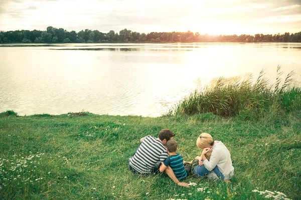 Fiatal gyönyörű szülők átölelve a fiatal fiúk, napnyugtakor a tónál. Családi séta a folyó mentén. — Stock Fotó
