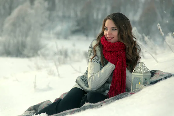 Красивая счастливая брюнетка в красном трикотажном шарфе сидит на одеяле на снегу зимой на фоне деревьев — стоковое фото