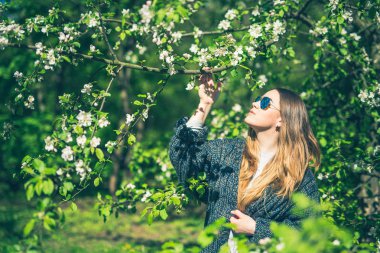 Mutlu genç kadın güneş gözlüğü ve bahçede bahar çiçek zevk örme ceket gülümseyen
