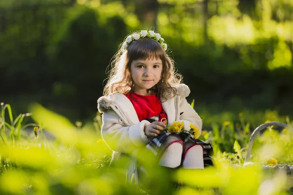 Nettes junges Mädchen trägt einen Löwenzahnkranz und lächelt, während es im Park auf Gras sitzt — Stockfoto