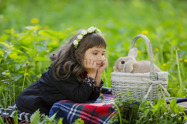 Uma menina em uma grinalda observa um coelho em uma cesta ao pôr do sol em um parque . — Fotografia de Stock