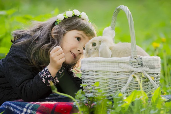 Uma menina em uma grinalda observa um coelho em uma cesta ao pôr do sol em um parque . — Fotografia de Stock