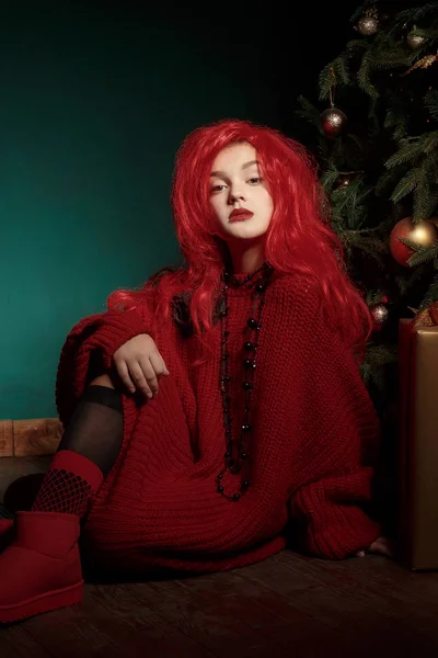 Ενός έφηβου κοριτσιού σε ένα κόκκινο πλεκτό πουλόβερ και περούκα κάθεται στο πάτωμα κοντά στο χριστουγεννιάτικο δέντρο και παρουσιάζει. Μόδας νέο έτος — Φωτογραφία Αρχείου
