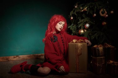 Kırmızı örgü kazak ve peruk bir genç kız bir Noel ağacı yakınındaki katta oturur ve sunar. Moda yeni yıl