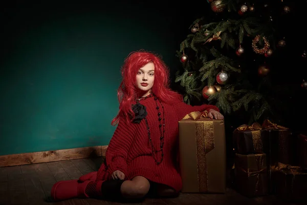 Девочка-подросток в красном трикотажном свитере и парике сидит на полу возле елки и дарит подарки. Новый год моды — стоковое фото