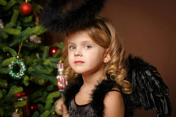 Nahaufnahme eines niedlichen kleinen blonden Mädchens mit blauen Augen in einem schwarzen Dämonenkostüm vor dem Hintergrund eines Weihnachtsbaums. neu — Stockfoto