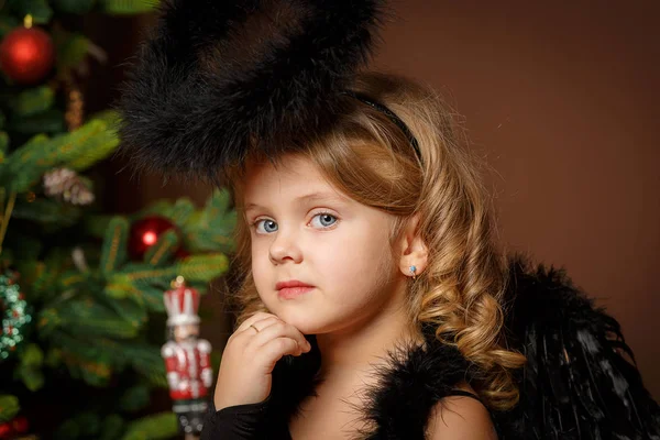 Nahaufnahme eines niedlichen kleinen blonden Mädchens mit blauen Augen in einem schwarzen Dämonenkostüm vor dem Hintergrund eines Weihnachtsbaums. neu — Stockfoto
